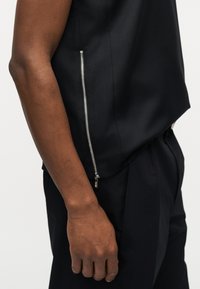 Άτομο με σκούρο χρώμα δέρματος που φοράει μαύρο αμάνικο τοπ με ορατό ασημί φερμουάρ στο πλάι και μαύρο παντελόνι που ταιριάζει, το χέρι χαλαρό δίπλα στο σώμα.