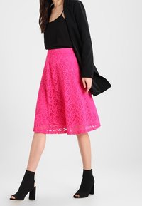 Nainen, joka on pukeutunut mustaan bleiseriin, mustaan hihattomaan paitaan, kirkkaanpinkkiin polven korkeuteen ulottuvaan pitsihameeseen ja mustiin avonaisiin korkokenkiin, seisoo valkoista taustaa vasten.