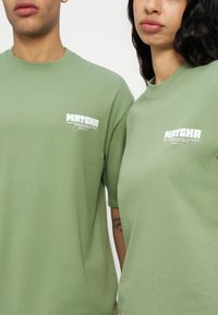 Δύο άτομα με ανοιχτοπράσινα μπλουζάκια που φέρουν λευκά λογότυπα με το κείμενο "MATCHA" στην πάνω αριστερή πλευρά, στέκονται δίπλα δίπλα με φόντο ένα λευκό τοίχο.