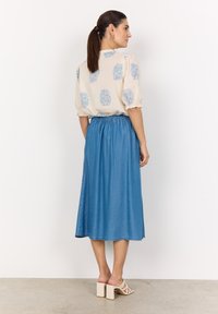 Blusa de un beige claro con patrones florales azules, de mangas cortas, y una falda maxi de mezclilla con cintura elástica, combinada con sandalias de tacón cuadrado en beige.