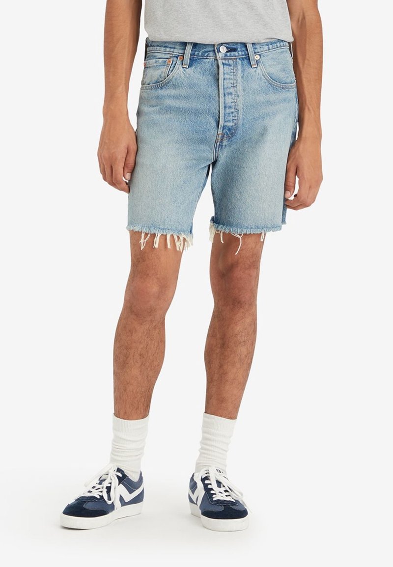 Shorts en denim bleu clair avec un ourlet effiloché, taille haute et lavage clair. Associés à des chaussettes blanches et des baskets bleues.