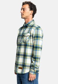 Koszula flanelowa z wzorem w kratę w odcieniach niebieskiego, żółtego i zielonego. Materiał wydaje się miękki, z kieszenią na piersi i metką markową na dolnym brzegu.