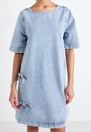 Femme portant une robe en jean bleu clair à manches courtes avec deux nœuds décoratifs près de l'ourlet sur un fond blanc uni.