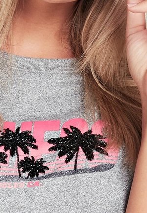 Gros plan sur une chemise grise avec des lettres roses et des motifs de palmiers noirs, portée par une personne aux cheveux blonds et longs.