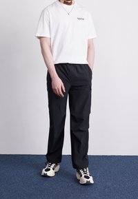 Man i vit Timberland-t-shirt, svarta cargobyxor och svart-vita sneakers som står på en blå matta mot en enfärgad vägg.