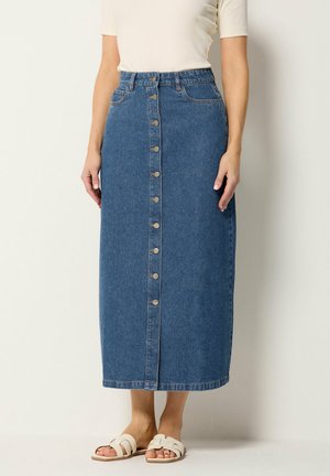 Mujer con una falda midi vaquera azul de talle alto con botones delanteros, top blanco de manga corta y sandalias blancas tipo deslizador.