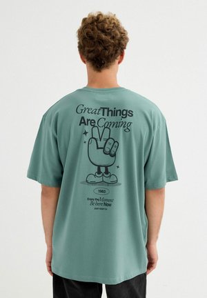 Persona con cabello rizado que lleva una camisa verde azulado mostrando una mano de dibujo animado haciendo el signo de la paz con el texto "Grandes cosas están por venir" en la parte trasera.