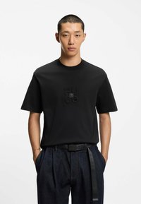 Camiseta de algodón negra con cuello redondo y mangas cortas. Presenta un logo texturizado de "HUGO" en el centro. Se lleva con pantalones de mezclilla oscuros.