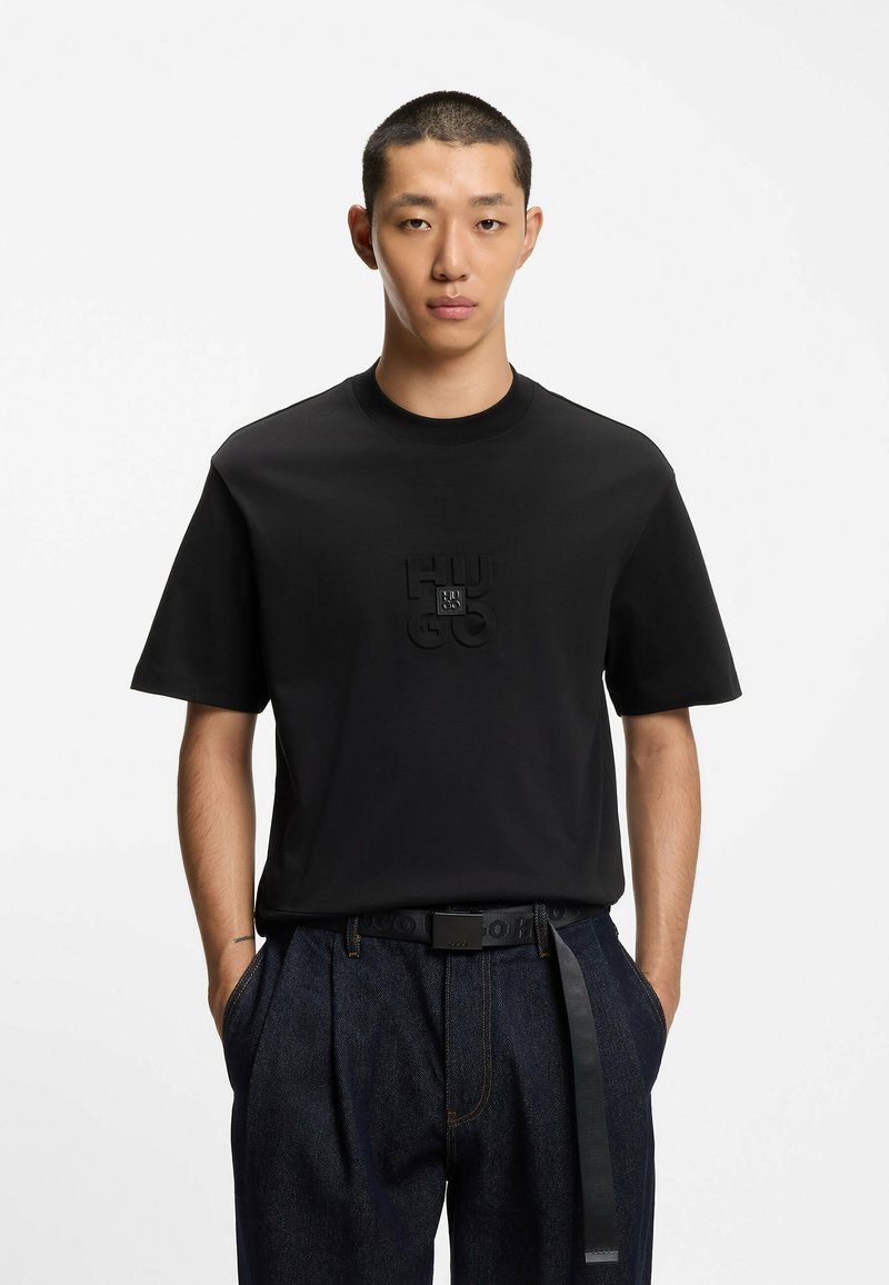 Camiseta de algodón negra con cuello redondo y mangas cortas. Presenta un logo texturizado de "HUGO" en el centro. Se lleva con pantalones de mezclilla oscuros.