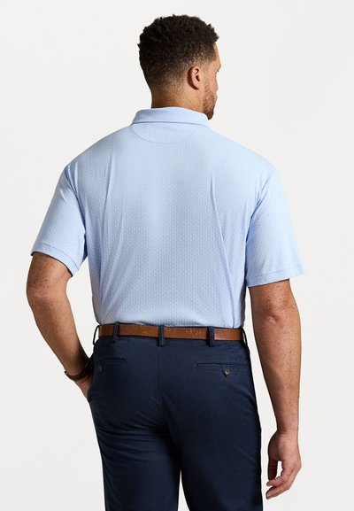 Homem a usar uma camisa polo de manga curta azul clara enfiada numa calça azul-marinho com cinto castanho, de costas contra um fundo branco liso.