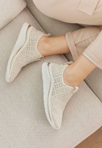 Leichte Slipper-Sneakers aus cremefarbenem Netzmaterial mit einer strukturierten Oberfläche und weißen Gummisohlen, die mit einem dezenten metallischen Akzent versehen sind.
