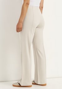 Osoba w beżowych spodniach z wysokim stanem i szerokich nogawkach oraz beżowych tenisówkach z brązowymi paskami, stojąca na tle jednolitego jasnego tła.