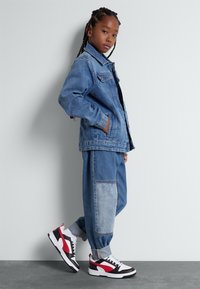 Denimjacka med avslappnad passform, ljusblå färg och framfickor, kombinerad med löst sittande tvåfärgade jeans och svart-röd-vit sneakers.