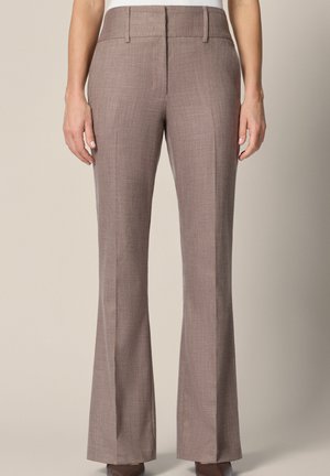 Kobieta ubrana w brązowe spodnie o prostych nogawkach, z wysokim stanem, stoi z ramionami swobodnie opuszczonymi wzdłuż ciała na neutralnym tle.