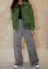 Zelená fleecová bunda s více kapsami, zipy a oranžovými akcenty. V kombinaci s volnými šedými kalhotami a černo-bílými teniskami.