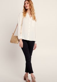 Chemise blanche à boutons avec un tissu texturé, manches longues et une poche poitrine, associée à un pantalon noir et des sandales à talons sangle beiges. Sac marron.