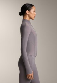 Fitted, dlhý rukáv, šedý top s vysokým golierom, s bezšvovým dizajnom a textúrovanou tkaninou. Nosený s odpovedajúcimi šedými legínami.