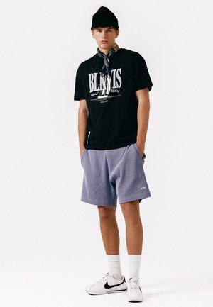 Ung mann som har på seg svart lue, svart grafisk t-skjorte, blå shorts, hvite sokker og hvite sneakers med svart Nike-swoosh, står med hendene i lomma.
