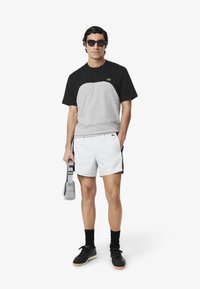 Camiseta de manga corta gris y negra, pantalones cortos blancos con franjas laterales negras, llevando una bolsa gris, usando gafas de sol, zapatos negros y calcetines negros.