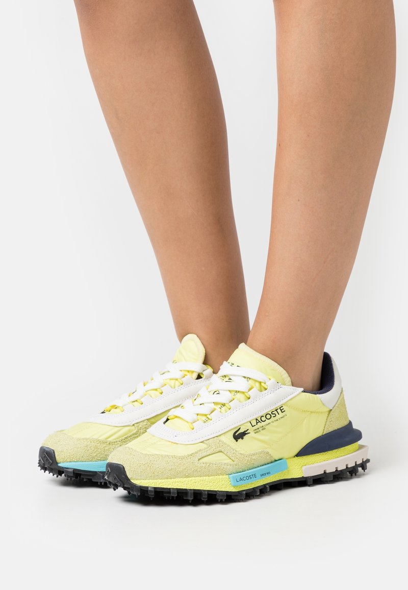 Lacoste Sneakers mit einem gelb-weißen Design, strukturiertem Wildleder und synthetischen Materialien, schwarzen Gummisohlen und leuchtend blauen Akzenten.