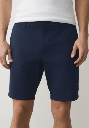 Mann trägt dunkelblaue Shorts und ein weißes T-Shirt, von der Taille bis zur Mitte des Oberschenkels gezeigt, steht mit entspannten Händen an den Seiten.