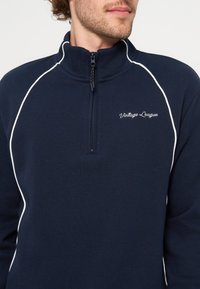 Homme portant un pull bleu marine à fermeture quart de zip avec des passepoils blancs et "Vintage League" brodé sur la poitrine.