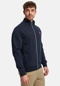Sweat-shirt zippé bleu marine avec un col montant, des poignets et un ourlet côtelés ; comporte un petit logo rouge sur la poitrine ; fabriqué en tissu doux.