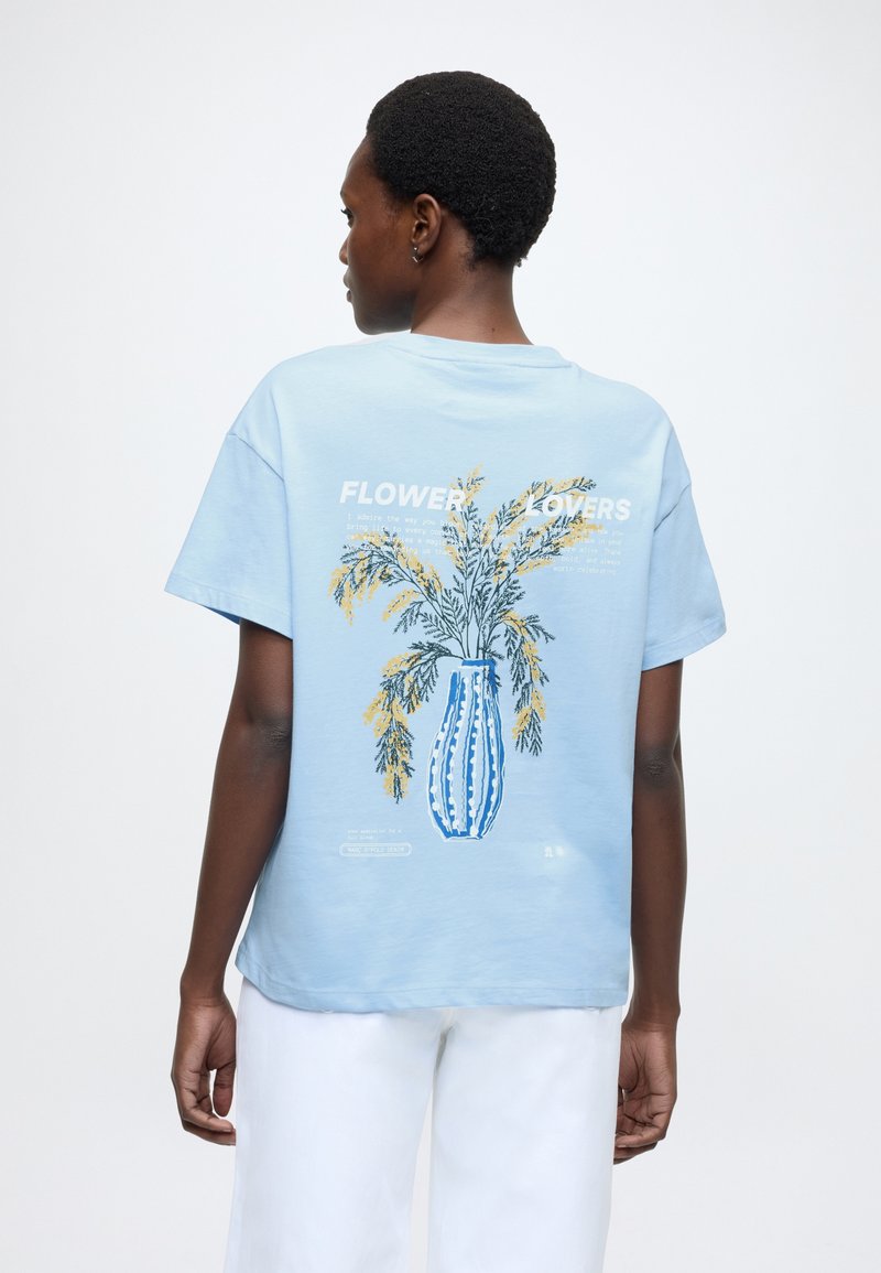 Camiseta de algodón azul claro con un gráfico de un jarrón que incluye flores amarillas y verdes. Mangas cortas, ajuste relajado y texto impreso en la parte trasera.