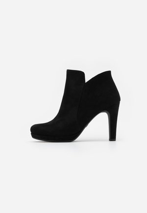 Bottine en daim noir avec un bout arrondi, un talon haut et un design découpé angulaire sur les côtés. Texture lisse et profil minimaliste.