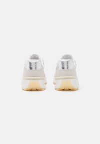 Light-colored sneakers with suede and metallic accents. Chunky sole with a textured pattern and visible logo on heel. Rounded design.