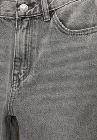 Gros plan d'un jean en denim gris montrant la poche avant, la boucle de ceinture, les coutures de la fermeture éclair et un bouton en métal avec marquage.