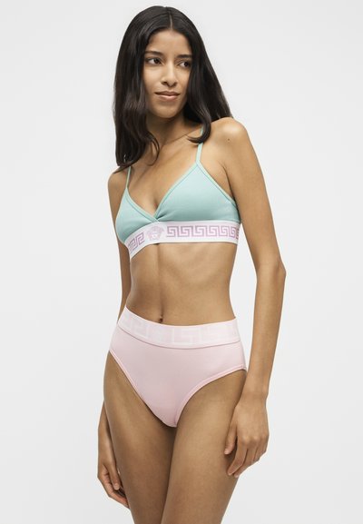 Γυναίκα που φοράει ένα ανοιχτό μπλε bralette και ανοιχτό ροζ εσώρουχο ψηλής μέσης, στέκεται μπροστά σε ένα απλό λευκό φόντο.