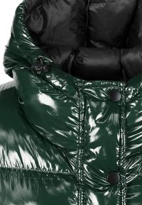 Zielona, błyszcząca kurtka puchowa z wysokim kołnierzem, czarnym wykończeniem i czarnymi zatrzaskami. Strukturalna powierzchnia z widocznymi detalami szwów.