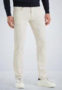 Jean slim coupe ajustée de couleur crème en denim. Présente des poches avant, un subtil emblème de la marque à l'arrière et un design épuré. Porté avec des baskets blanches.