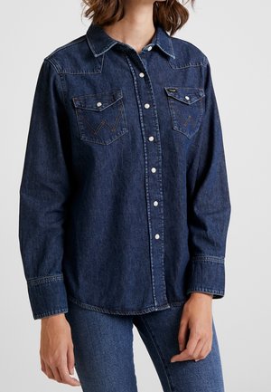 Kvinde iført en mørkeblå denimknap-op-skjorte med to brystlommer og lyse trykknapper, kombineret med blå jeans.