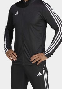 Chaqueta deportiva negra de manga larga con cremallera frontal y detalles de tres rayas blancas en las mangas, combinada con pantalones a juego. Presenta el logo de adidas.