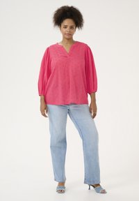 Blouse fuchsia avec motif texturé et manches courtes, associée à un jean droit bleu clair et des sandales à talons ouvertes assorties.