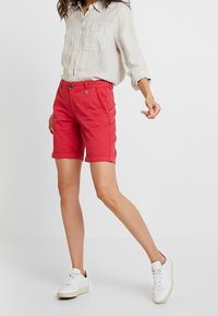 Pantalones cortos de algodón rojos con dobladillo enrollado, con bolsillos laterales, cierre de botón en la parte delantera y un diseño de frente plano. Acompañados de una camisa de lino ligera.