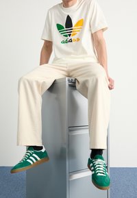 Krémové tričko s černým, zeleným a žlutým logem Adidas; béžové volné kalhoty; zelené a bílé semišové sneakers; sedí na stříbrném zásuvkovém kabinetu.