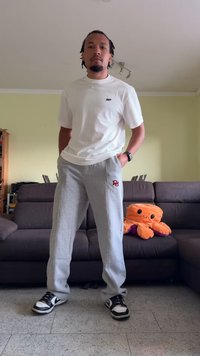 T-shirt de manga curta branca, calças de fato cinzentas com logótipo vermelho e sapatilhas pretas e brancas. De pé numa sala de estar com um brinquedo de peluche.