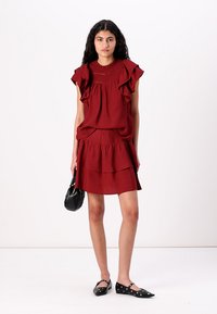 Red sleeveless dress with ruffled shoulder accents, tiered skirt, and textured bodice. Paired with black flat shoes and a small handbag.