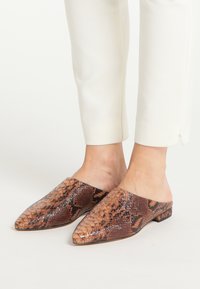 Bruine mules met slangenprint, een puntige neus en een platte hak. Geverfde lederen oppervlakte met glanzende accenten. Gecombineerd met strakke witte broeken.
