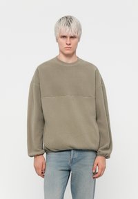 Olivenfarget fleecegenser med rund hals og ribbestrikkede mansjetter, med et kontrasterende horisontalt panel på brystet. Brukes sammen med lyse jeans.
