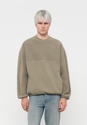 Olivenfarget fleecegenser med rund hals og ribbestrikkede mansjetter, med et kontrasterende horisontalt panel på brystet. Brukes sammen med lyse jeans.