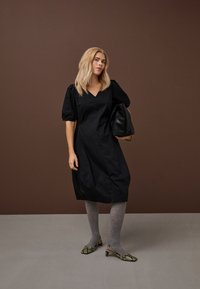 Ženska s svetlo blond lasmi nosi črno obleko z napihanimi rokavi, sive nogavice, salonarje z živalskim vzorcem na peti in drži veliko črno teksturirano torbico na rjavi podlagi.