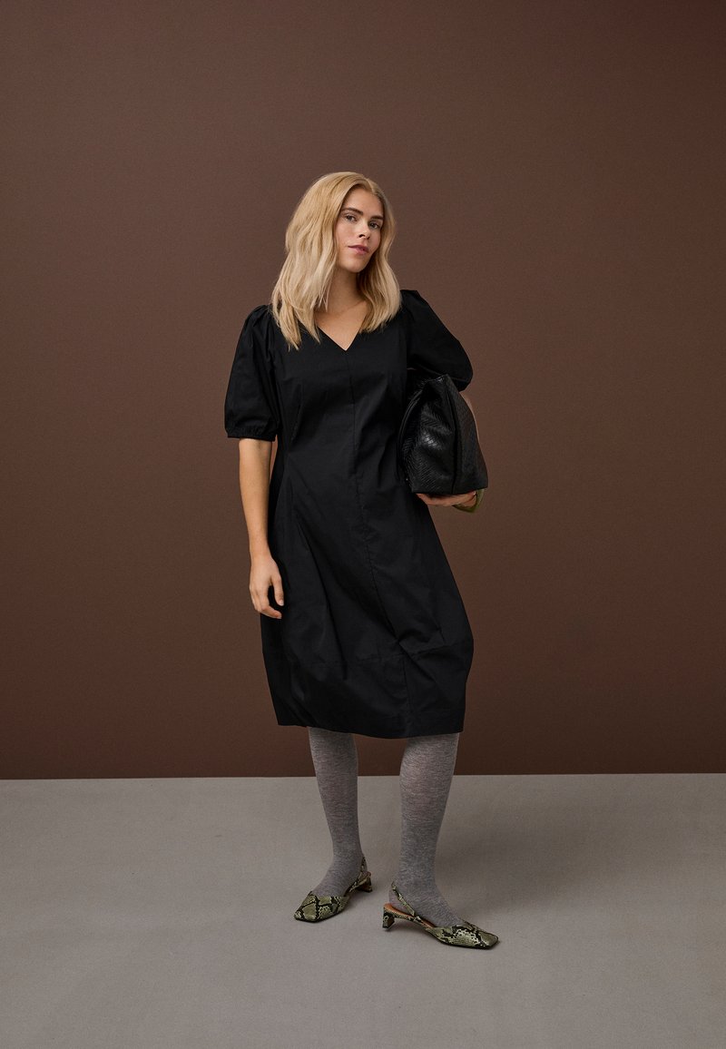 Ženska s svetlo blond lasmi nosi črno obleko z napihanimi rokavi, sive nogavice, salonarje z živalskim vzorcem na peti in drži veliko črno teksturirano torbico na rjavi podlagi.