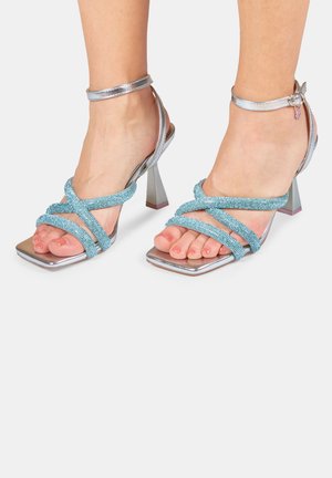 Sandali argentati con tacco a forma quadrata arrotondata, dotati di cinturini incrociati turchesi decorati con piccole perline e una cinghia metallica alla caviglia.