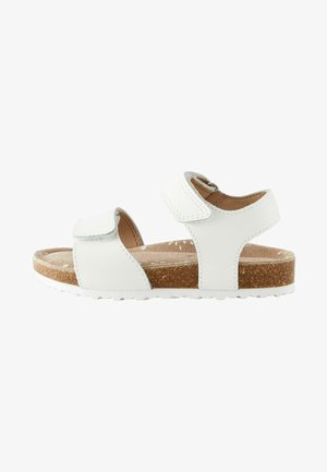 White sandal with adjustable velcro straps, cork sole, and textured footbed. Features a flat design and durable rubber outsole for grip.