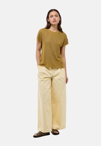 Femme debout avec les mains dans les poches portant un haut à manches courtes moutarde, un large pantalon crème et des sandales brun foncé sur fond blanc.