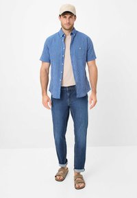 Camicia blu e bianca a righe con maniche corte, t-shirt beige a girocollo, jeans in denim scuro e sandali a scivolo marroni. Outfit casual.